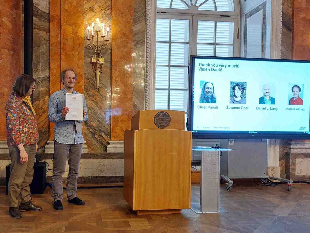 Two people stand by a podium; one holds a certificate. A screen displays four names and photos.